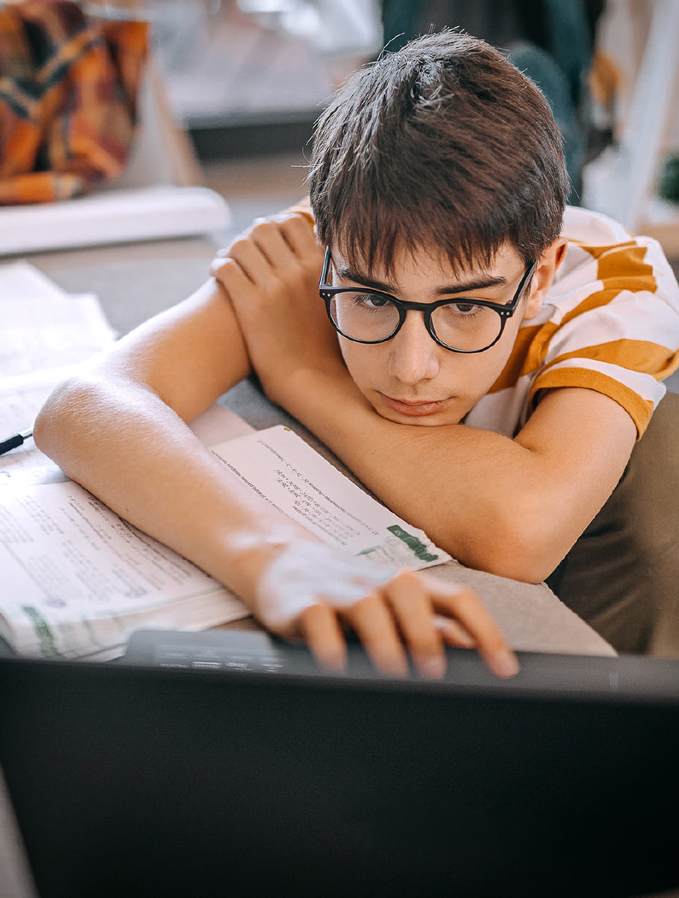 A boy wearing glasses resting on his textbooks in front of a laptop